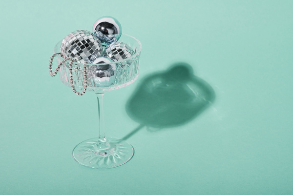 Small Christmas balls of silver color and in the form of a disco ball and beads lie in a champagne glass on a high leg casting a shadow on a light blue background