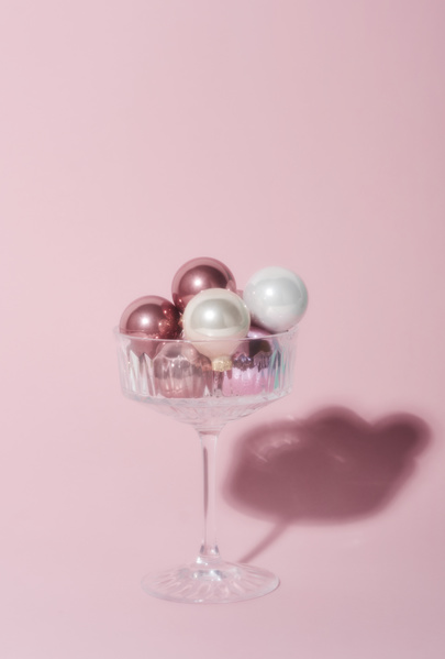 Blurred image of a champagne glass on a high leg with small pink and white Christmas balls on a light-colored background