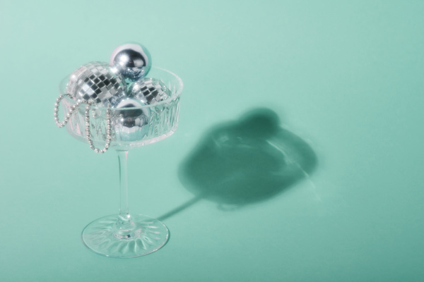 Blurred photo of a champagne glass filled with silver Christmas balls and standing against a light blue background casting a shadow