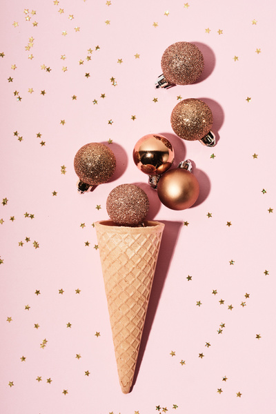 Golden Christmas balls with different surfaces lie above the waffle cone resembling ice cream on a light pink background strewn with yellow stars
