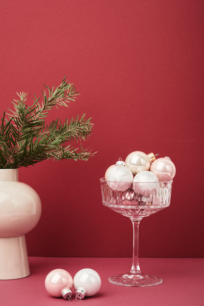 Small white glossy Christmas balls lie in a wide glass next to which there are fir tree branches in a vase and two balls lie