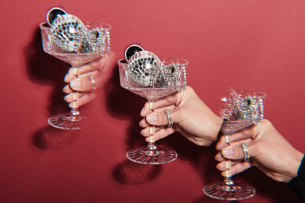 Repeated images of a champagne glass filled with small silver Christmas tree balls and beads in a womans hand with a ring on a fuchsia background