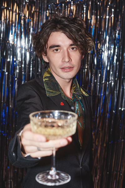 Portrait of a young brown-eyed man with dark curly hair and dressed smartly holding out a glass of champagne against the background of lametta