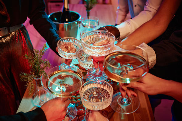 A lot of champagne glasses in the hands of people clinking them over a festive table made of wood decorated with Christmas tree branches in a vase