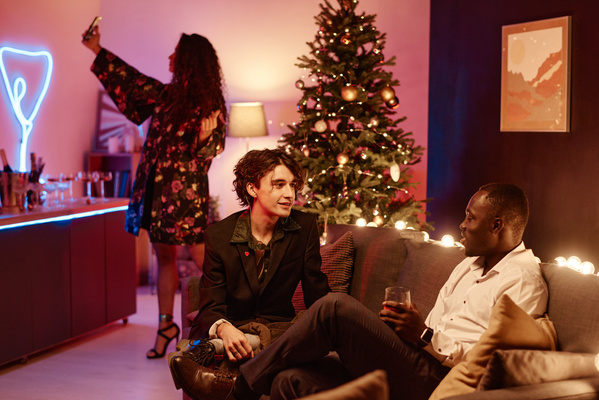 Two dark-haired friends in elegant outfits sitting on the sofa talking in a cozy room with a Christmas tree decorated with balls and a garland