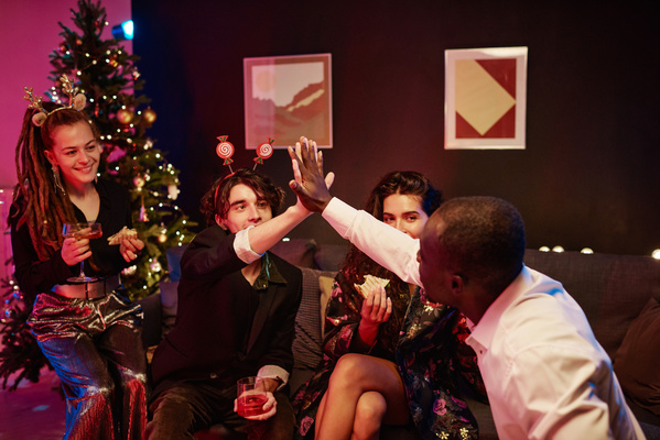 Young men from a group of friends gathered to celebrate Christmas give each other a high five sitting on the couch with beautifully dressed womans in a room with a Christmas tree