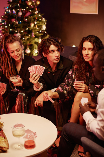 A group of familiars gathered to celebrate Christmas sitting on the sofa at a table with drinks in glasses and playing cards in a cozy room with a Christmas tree