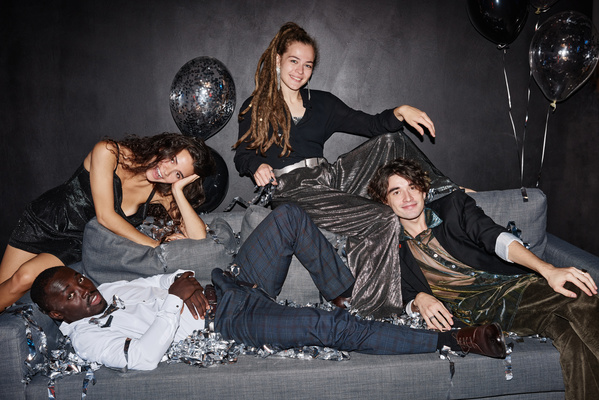 Young friends in dark festive outfits smiling pose for a photo sitting on the sofa and its back with silver confetti and baloons against a dark wall