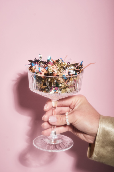 A blurred photo of a champagne glass on a high stem full of shining confetti in a female hand with a pearl ring on a pink background