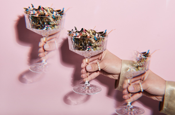 A repeating image of a champagne glass on a high leg with shimmering confetti in a female hand with a pearl ring on a pink background