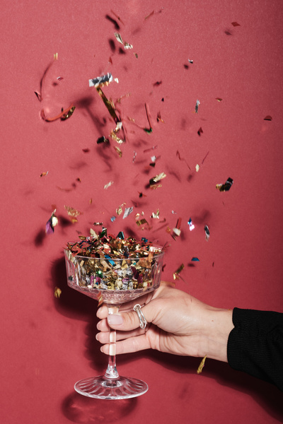 A wide champagne glass on a high leg with fluttering confetti in a female hand with a silver ring on her thumb on a fuchsia background
