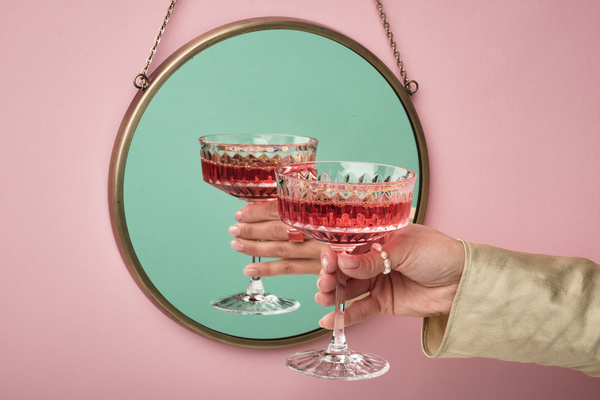 A faceted wide glass filled with pink drink in a womans hand with a ring of pearl beads on her thumb and a light sleeve is reflected in a round mirror hanging on a chain on a pink wall