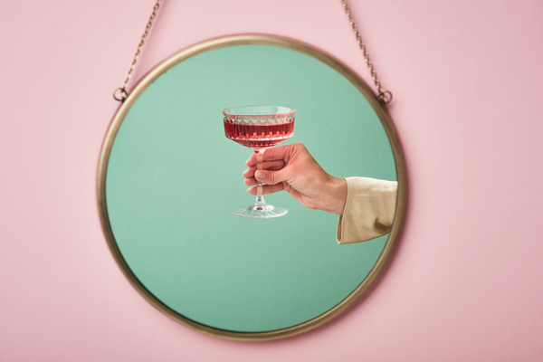 Reflection of a glass with pink champagne on a high leg in a womans hand with a light sleeve on a blue background in a round mirror that is hang on a chain on a pink wall