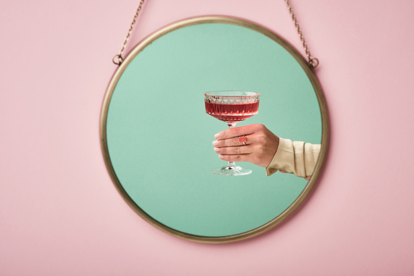 Reflection of a faceted glass with a pink drink in a female hand with a light sleeve and a ring with a rectangular stone on a blue background in a round mirror that is hung on a chain on a pink wall