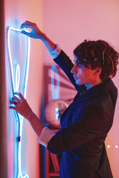 A young man with curly hair dressed in a dark jacket hangs a neon decoration in the form of a glass on the wall