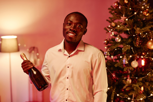 A man in a white shirt smiling and holding a bottle of champagne in his hand stands in a cozy room with pink lighting against the background of a decorated Christmas tree
