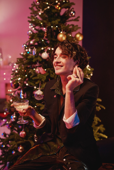 A man with curly hair in a dark outfit smiling and holding a glass of champagne in his hand sitting in a cozy room with pink lighting with a decorated Christmas tree behind