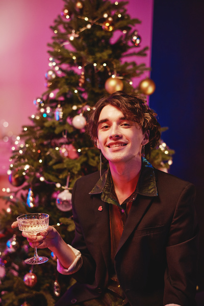 A young man with dark curly hair in a dark outfit smiling and holding a glass of champagne in his hand sitting in a cozy room with pink lighting with a decorated Christmas tree behind