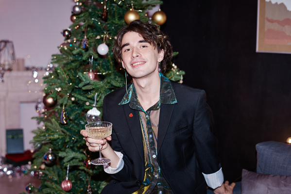 A guy with dark curly hair in smart clothes smiling and holding a glass of champagne in his hand sitting in a cozy room with a decorated Christmas tree behind