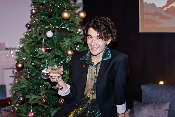 A cute dark-haired man in an eccentric outfit holding a glass of champagne in his hand and laughing sits against the background of a Christmas tree