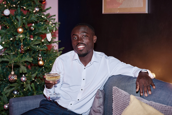 A guy with a beard in a white shirt and dark blue trousers sitting on the sofa with a glass of champagne with his arm thrown over the back with a Christmas tree behind