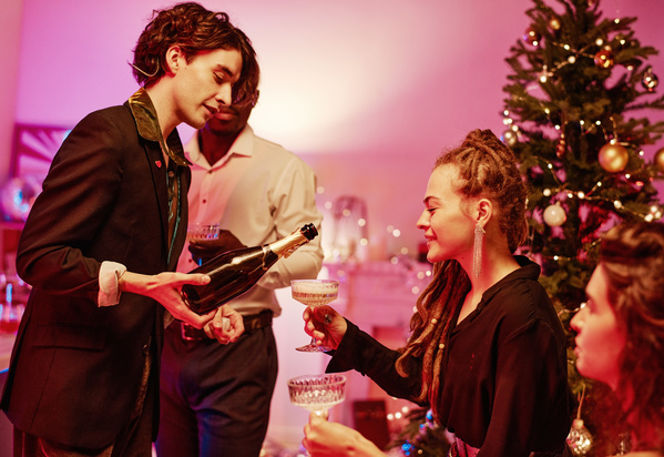 A young handsome man with curly hair in a smart suit pours champagne to his womanfriend with dreadlocks at a Christmas celebration