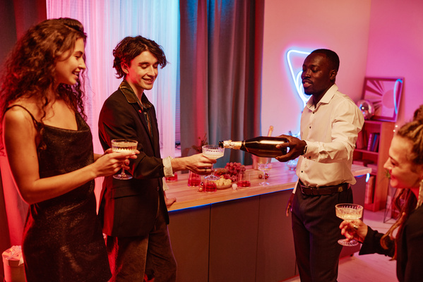 A guy in a white shirt pours champagne from a bottle to a friend in a faceted glass on a high leg in a room with pink lighting and young women with glasses