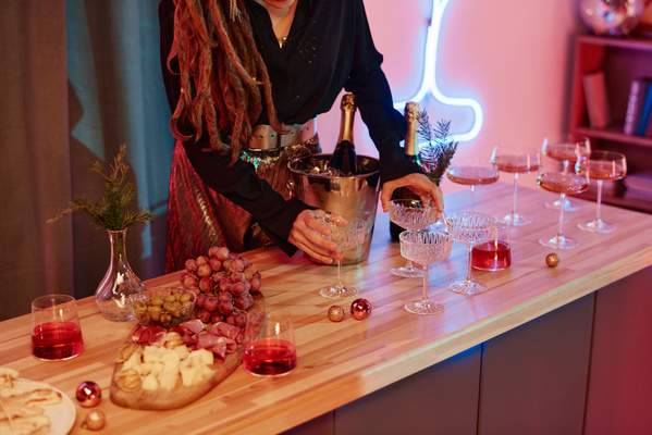 A woman with dreadlocks dressed in an elegant outfit places glasses on a festive table decorated with Christmas attributes with cheese and meat plateaus and bottles of champagne