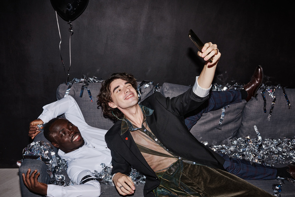 A curly-haired guy in an extravagant outfit takes a selfie with a friend in a white shirt lying on the couch strewn with silver confetti