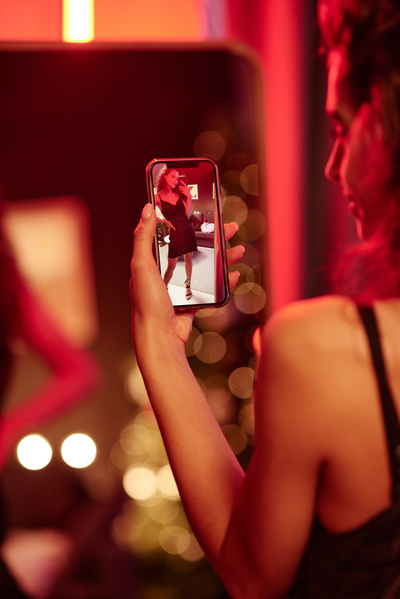 A woman with long hair in a black short dress takes a photo in the mirror at a Christmas party in a room with a dim pink light
