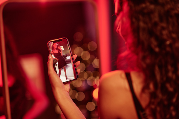 A woman with curly hair in a black short dress takes pictures of herself in the mirror posing at a Christmas party in a room with a dim pink light