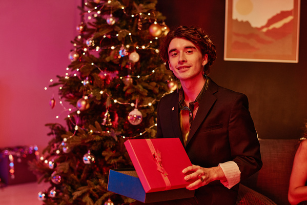 A young man with dark curly hair dressed in a black jacket and a shining shirt sits against the background of a Christmas tree and opens a gift box