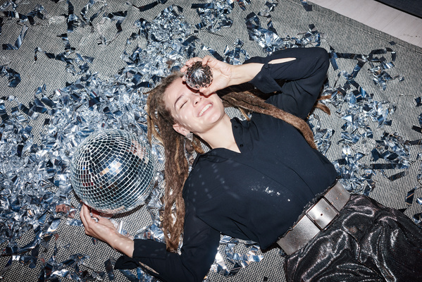 A woman with dreadlocks dressed in a dark outfit lies on a blanket strewn with silver confetti with a disco ball and smiling to the teeth puts a Christmas ball to her face