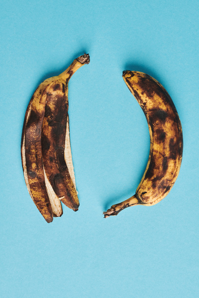 A ripe banana with dark spots lying with the stem down with the peel of a ripe banana on a blue surface