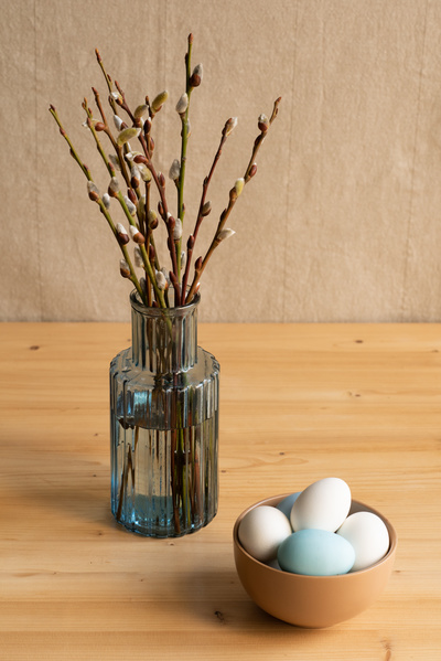 A brown bowl with Easter eggs and a blue vase with willow twigs on a wooden table