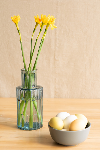 A grey bowl with Easter eggs and a blue vase with a bouquet of yellow daffodils on a wooden table