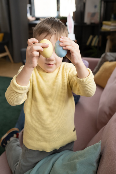 A blond boy in a yellow sweater holding yellow and blue Easter eggs kneeling on a pink sofa