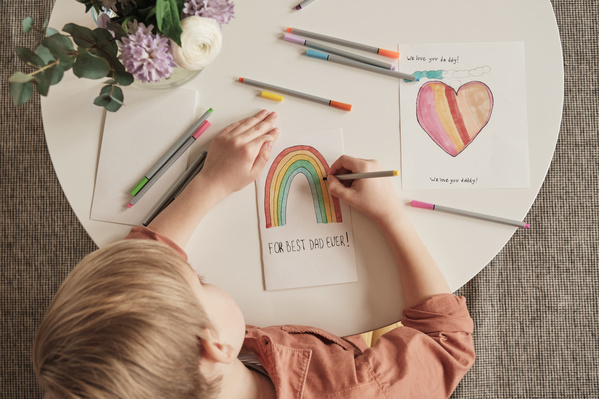 A blond-haired boy sitting at a table coloring a rainbow with markers on a handmade postcard for Fathers Day