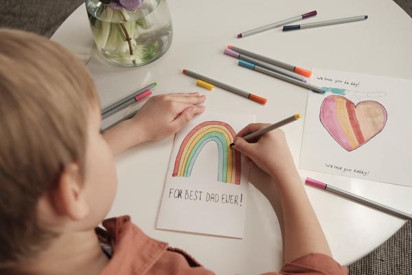 A blond boy sitting at a table painting a rainbow with markers on a homemade Fathers Day card