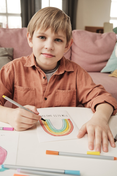 A blond boy in a brick-colored shirt drawing a rainbow on a homemade Fathers Day card