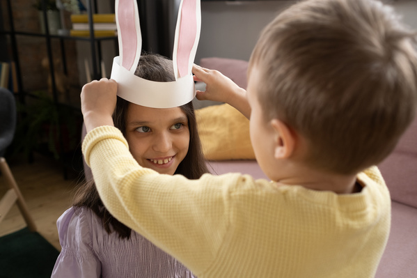 A blond boy puts bunny ears headband made of paper on his smiling sister