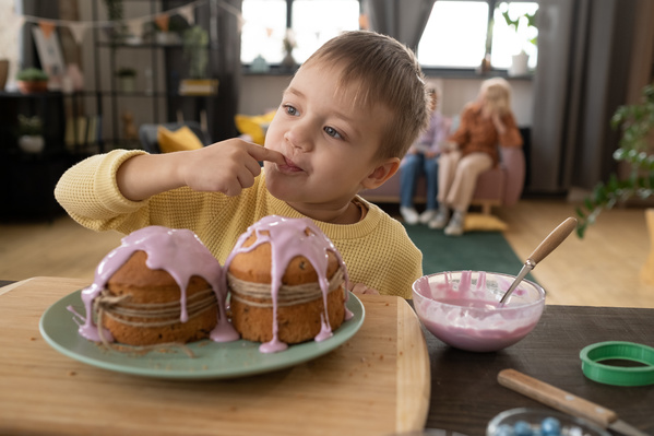 A blond boy in a yellow sweater tasting pink icing for traditional Easter cakes standing on the table and decorated with twine