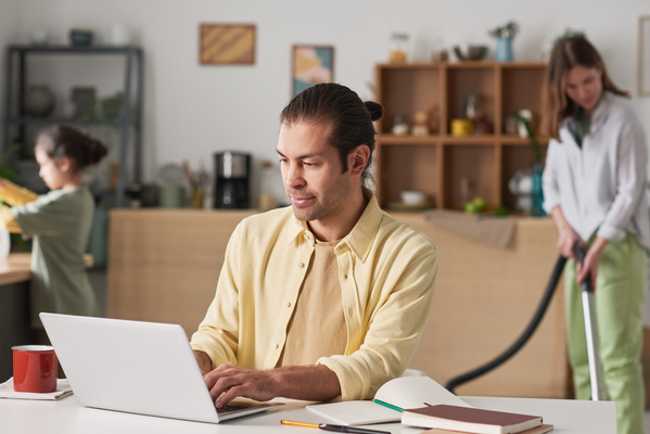 A man in a yellow shirt over a T-shirt is focused on working at the laptop while sitting at the desk