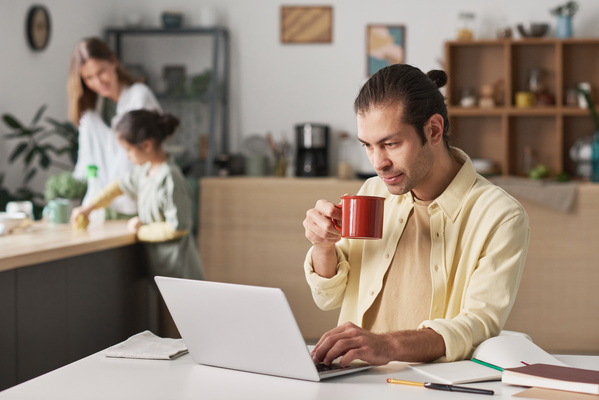 A busy man in yellow clothes sitting holding a red mug at a desk with a laptop on it