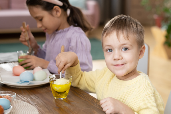 A smiling boy with blond hair in a yellow sweater dipping an egg in yellow dye sitting at the table with his sister