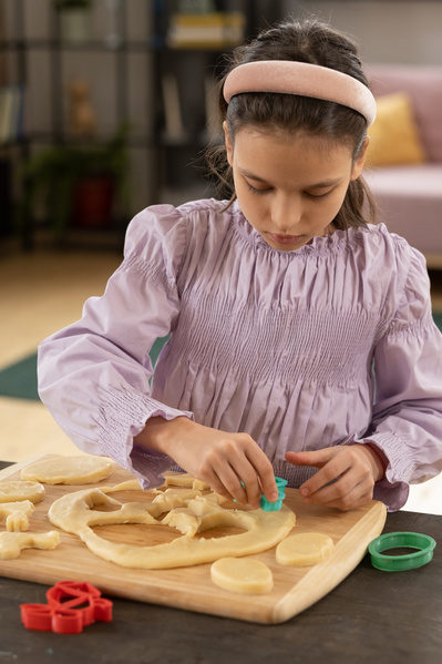 A girl in a pink blouse cutting cookies out of dough on a wooden cutting board with colored plastic molds