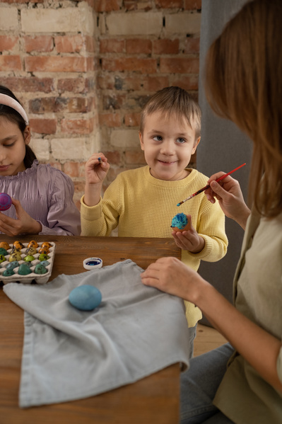 A mother helping her son in a yellow sweater to color an Easter egg blue with a brush