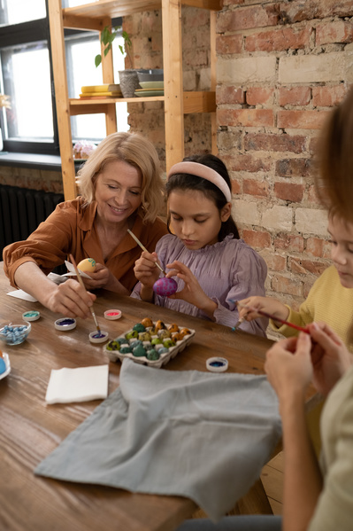 A girl in a pink blouse and a grandmother paint Easter eggs with paints sitting at a wooden table
