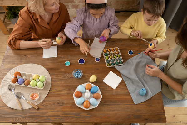 A family consisting of two children a mother and a grandmother painting Easter eggs sitting at a wooden table on which materials for this are laid out