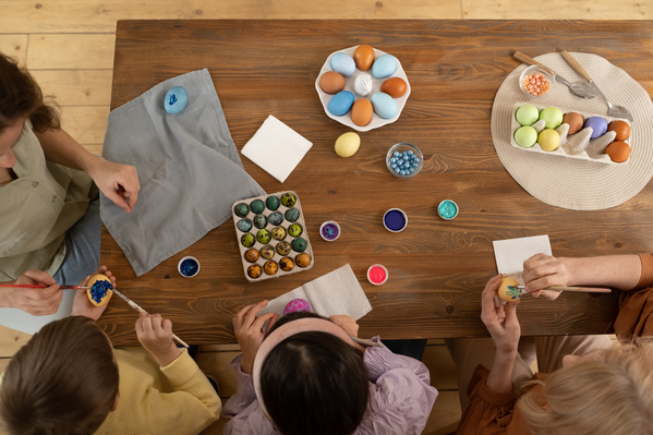 Top view of a family consisting of two children a mother and a grandmother coloring Easter eggs and sitting at a wooden table on which materials for this are laid out